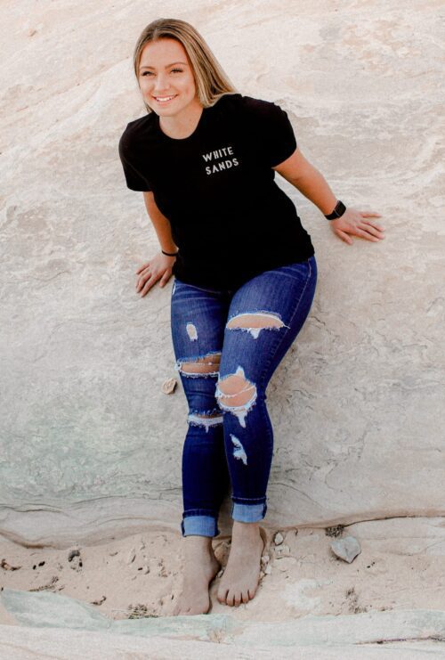 Female leaning against a rock with a white sands national park t-shirt and khaki pants
