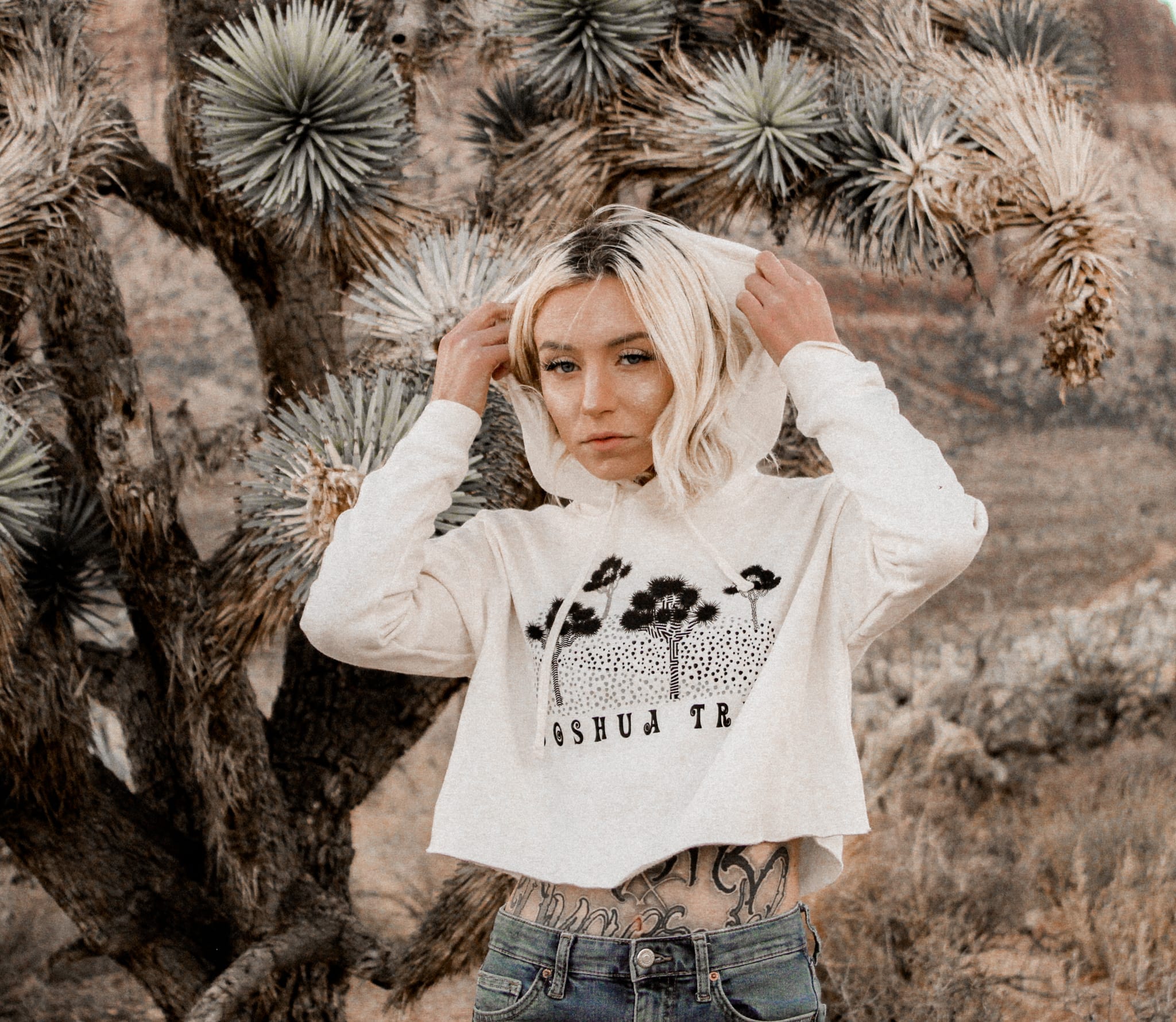 female standing next to a joshua tree with mountains in the background wearing a joshua tree hoodie