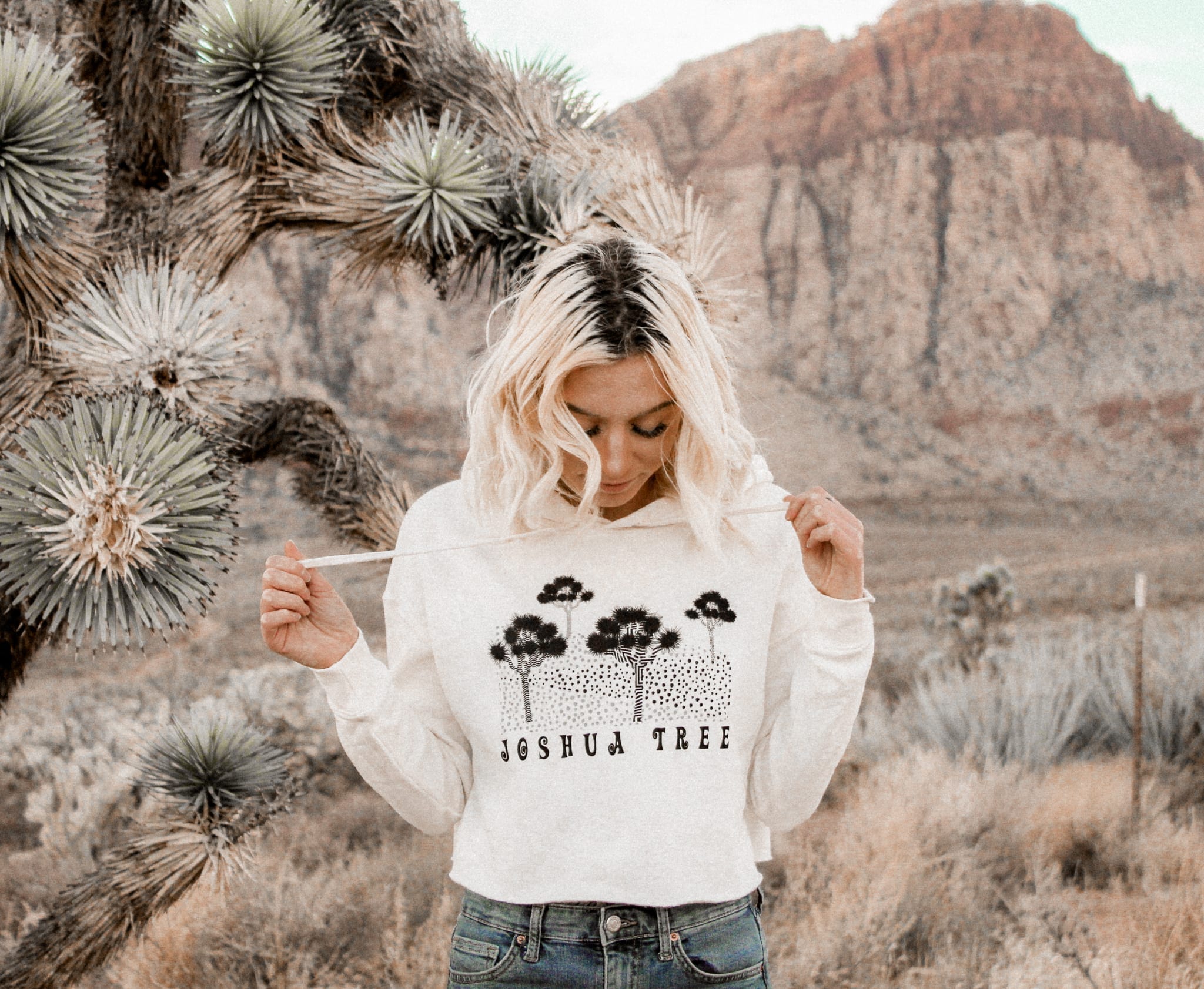 female standing next to a joshua tree with mountains in the background wearing a joshua tree hoodie